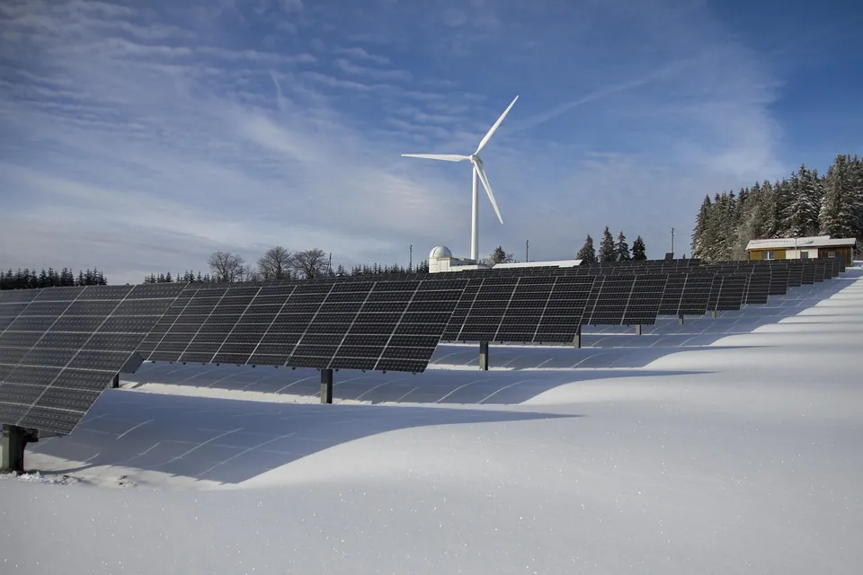 Rangées de panneaux solaires dans un paysage enneigé, avec une éolienne et des sapins en arrière-plan