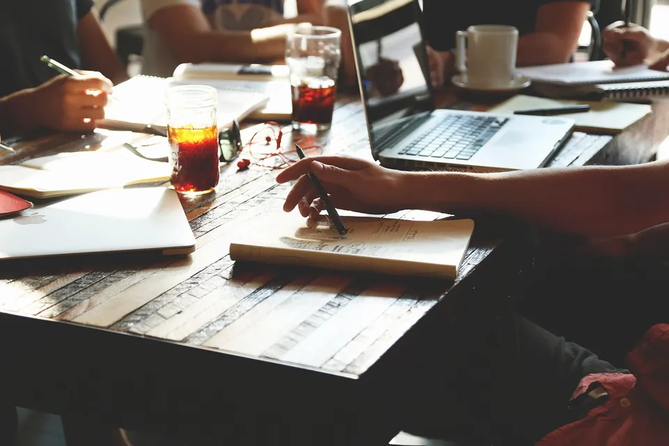 Personnes assises autour d'une table en bois, carnets et ordinateur ouverts, en pleine discussion