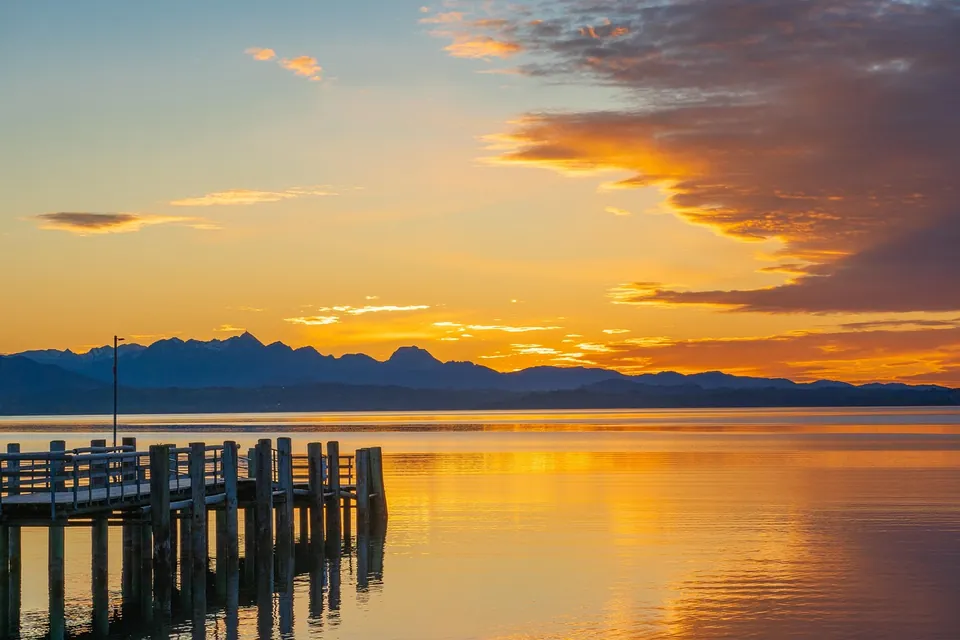 Coucher de soleil orangé sur un lac, avec un ponton en bois et une chaîne de montagnes à l'horizon